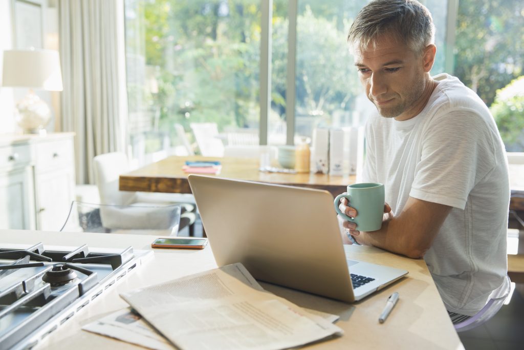 Man drinking coffee and working at laptop in kitchen.