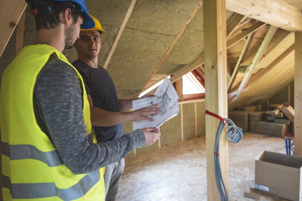 Construction workers reviewing blueprint at construction site. Roof with mineral rockwool in wall section. House under construction with insulation glass wool on an attic floor.