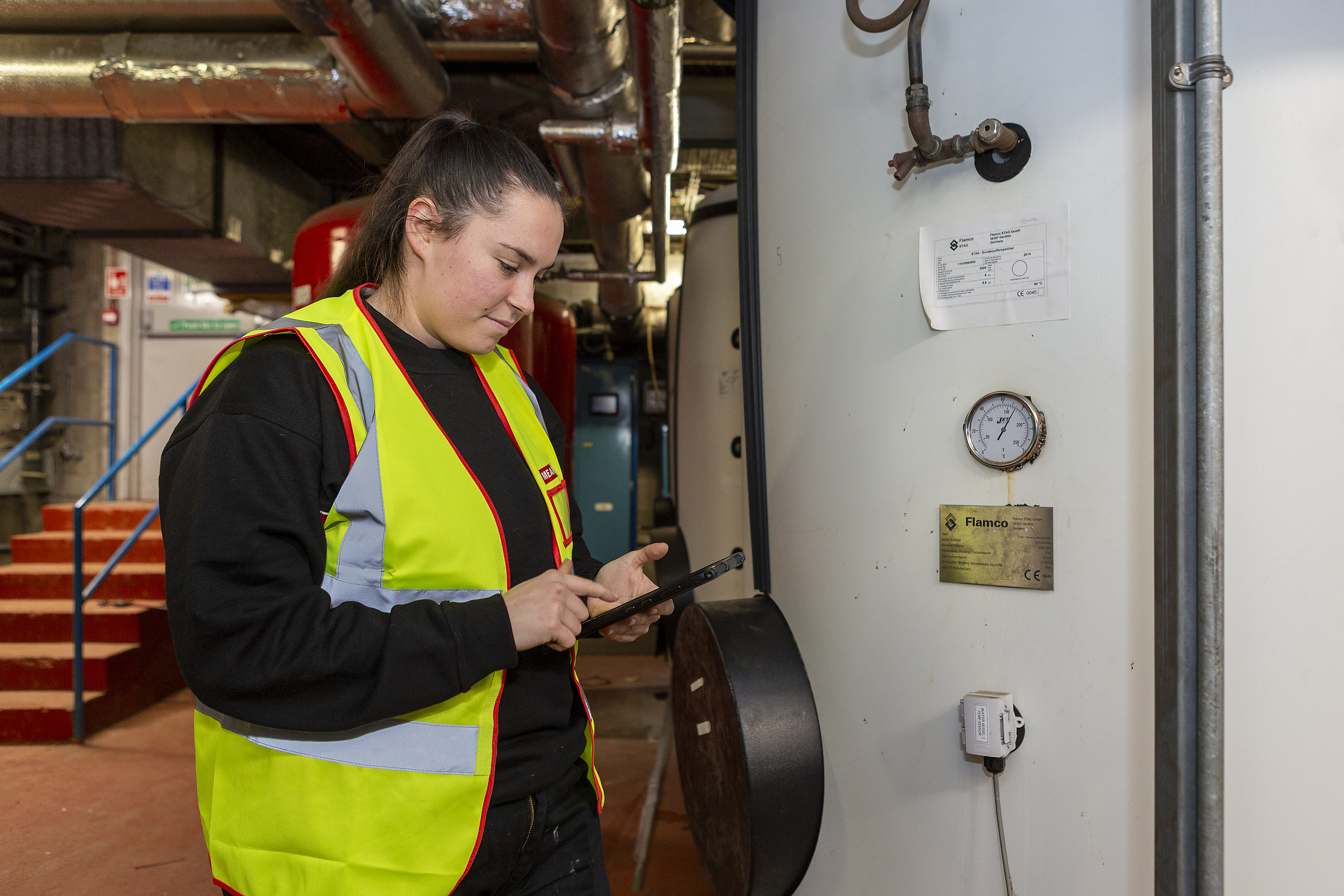A person wearing a high‑visibility vest inspects industrial heating equipment in a plant room, holding a tablet while checking gauges and pipes on a large cylindrical unit.