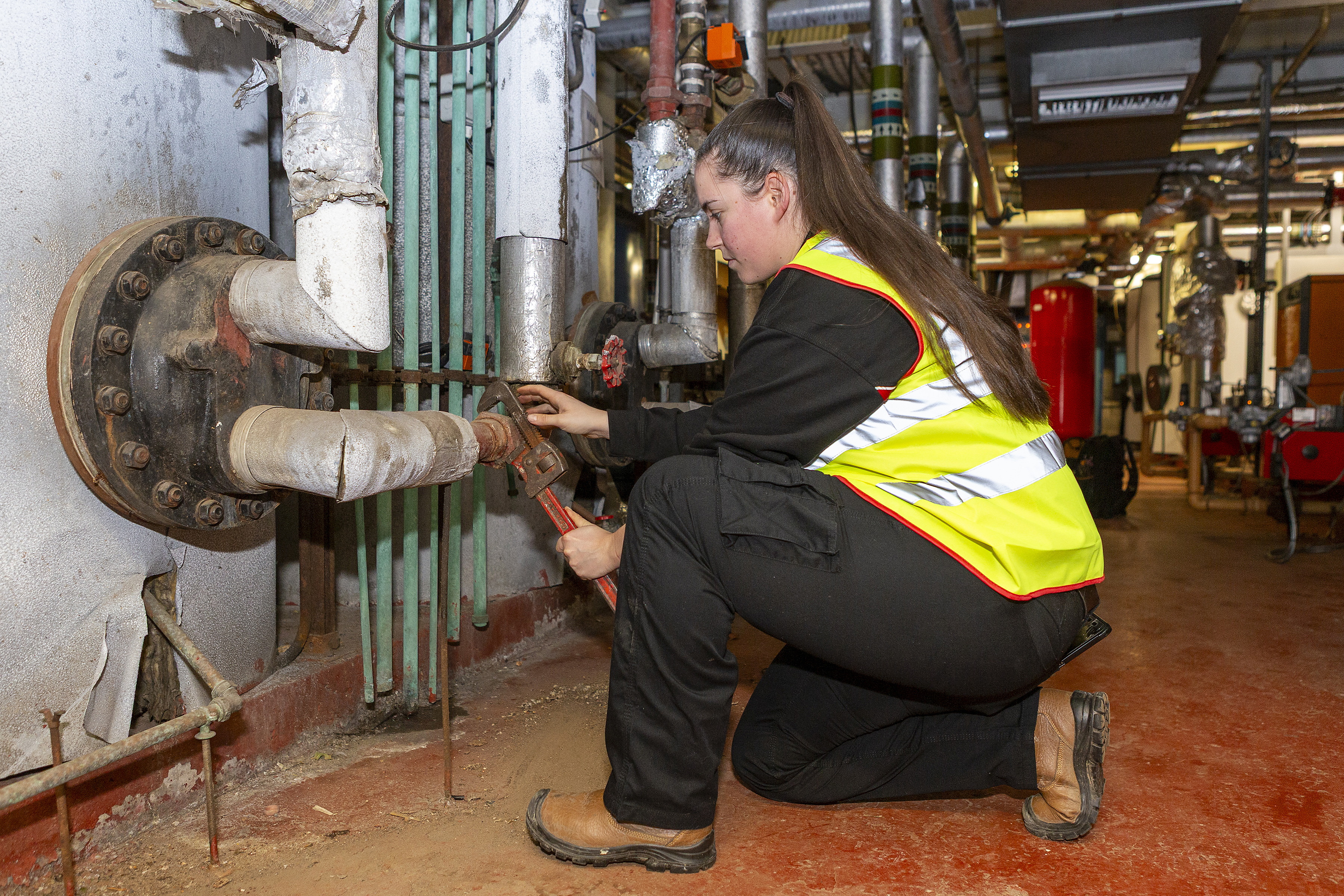 A worker in a high-visibility vest kneels in a plant room, tightening a pipe valve with a wrench among industrial piping.
