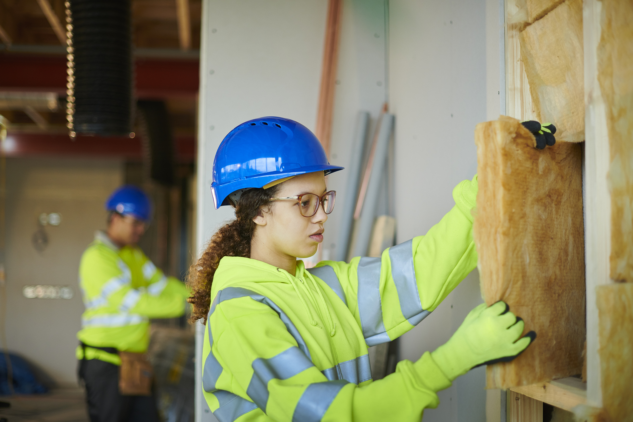 A building site where a worker wearing a blue hard hat, high‑visibility jacket and gloves is fitting insulation into a wall cavity. Another worker in similar safety clothing is visible in the background. The wall framing and insulation materials are clearly visible indoors.